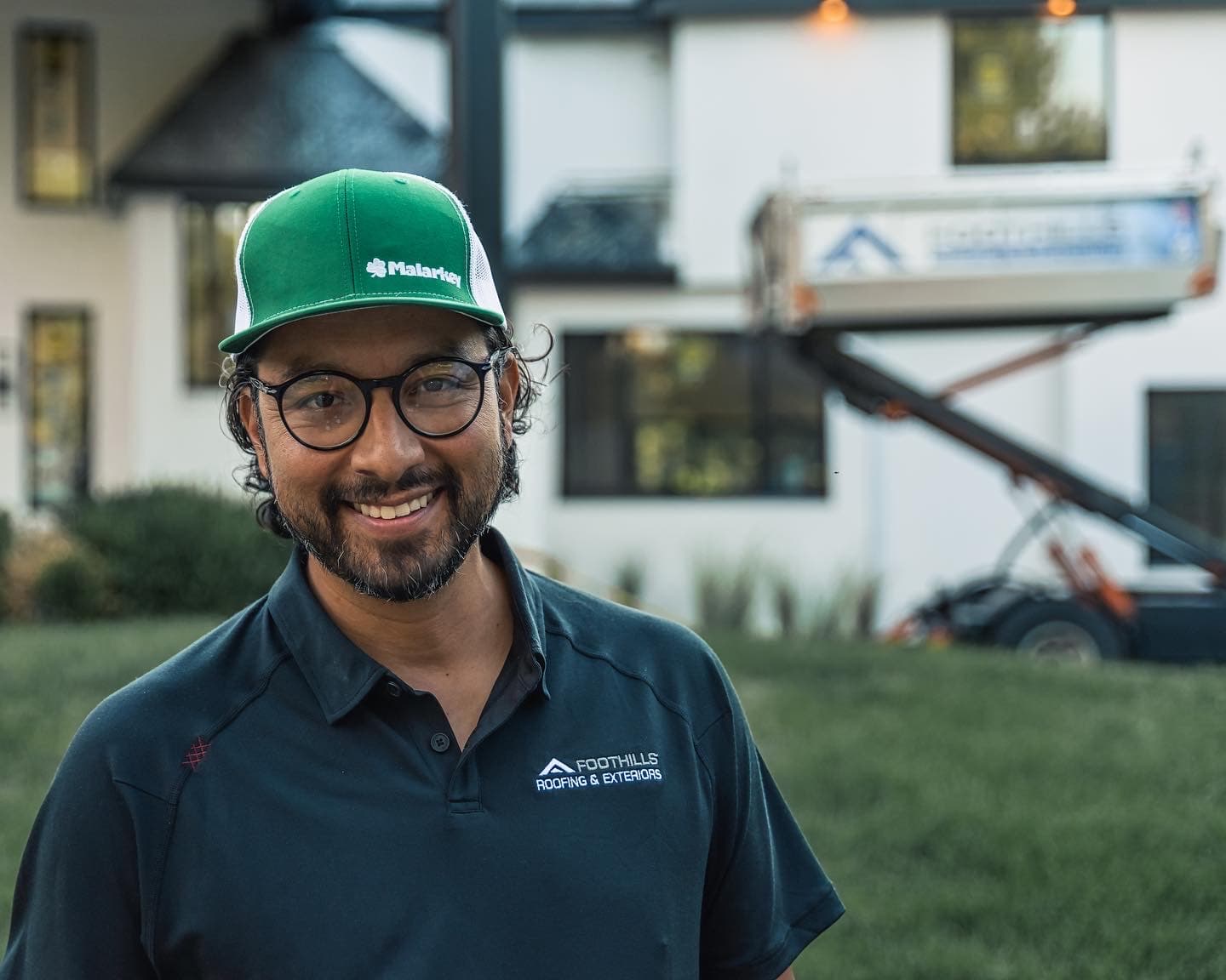 A smiling Foothills team member in a green Malarkey cap and a Foothills Roofing & Exteriors polo, with the company lift parked behind